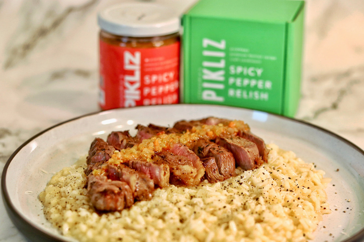 Creamy parmesan risotto topped with sliced butter-basted steak and Alexandra’s Pikliz® relish, with Pikliz products in the background.