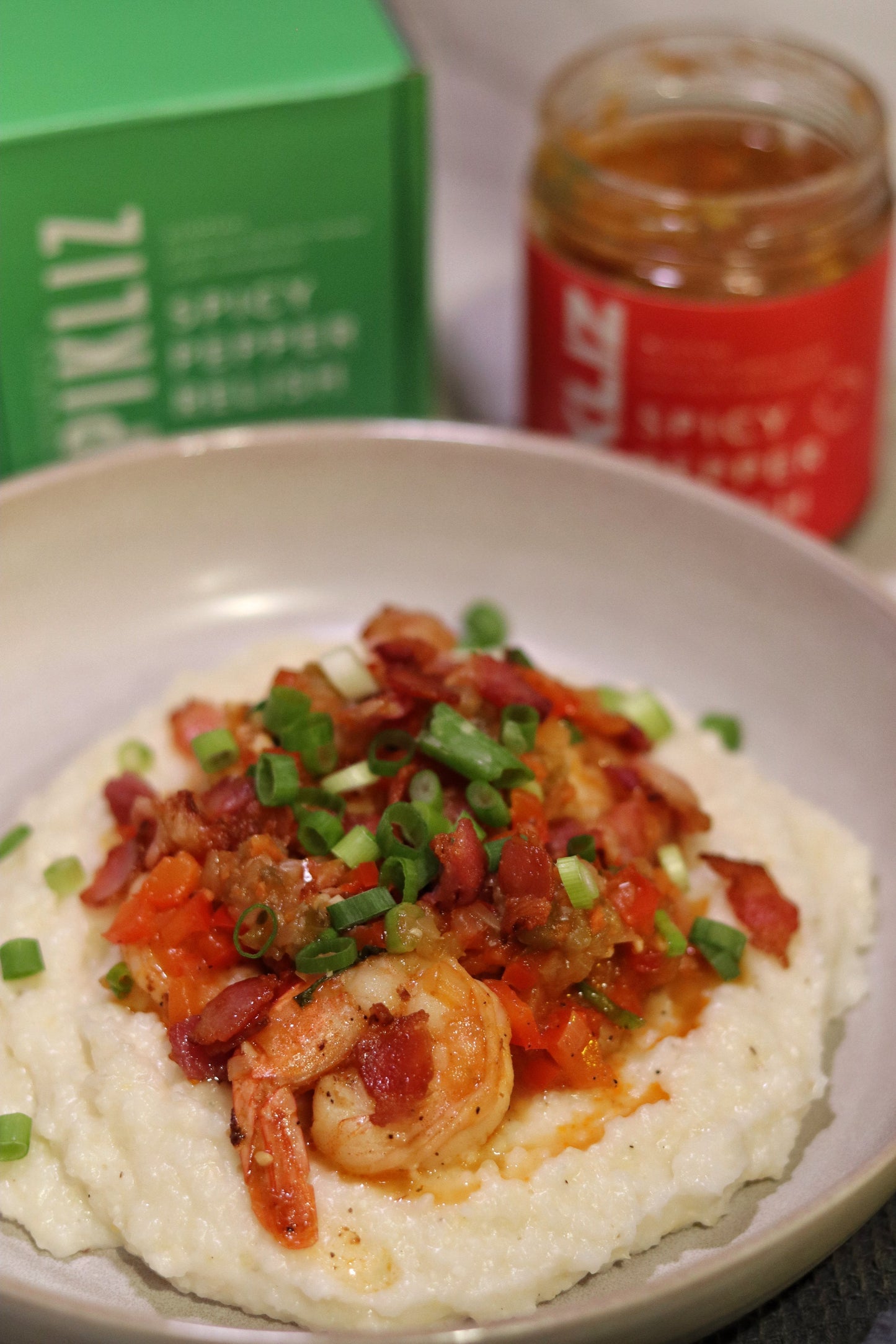Cajun shrimp and grits topped with Alexandra’s Pikliz Spicy Pepper Relish, crispy bacon, green onion, and creamy cheese grits